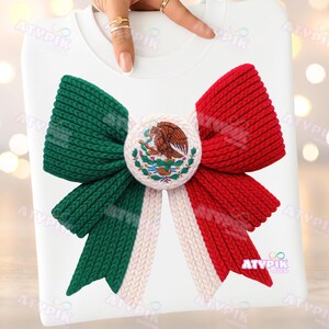 May include: White sweatshirt with a large bow design in the colours of the Mexican flag: green, white, and red. The bow has a central emblem with the Mexican coat of arms. The design is made of a knitted material.