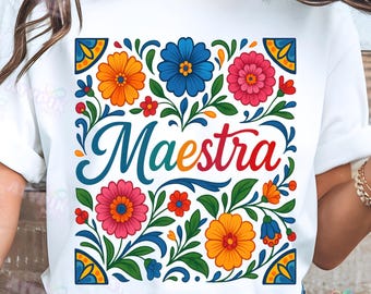 May include: White t-shirt featuring a vibrant floral design with orange, blue, and pink flowers, green leaves, and the word "Maestra" in colorful script. The design has a folk art style with decorative corner elements.