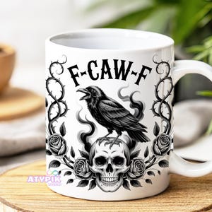 May include: White ceramic mug with a black raven design. The raven is perched on a skull, surrounded by roses and thorny vines. The mug has the text "F-CAW-F". The design is in black and white.