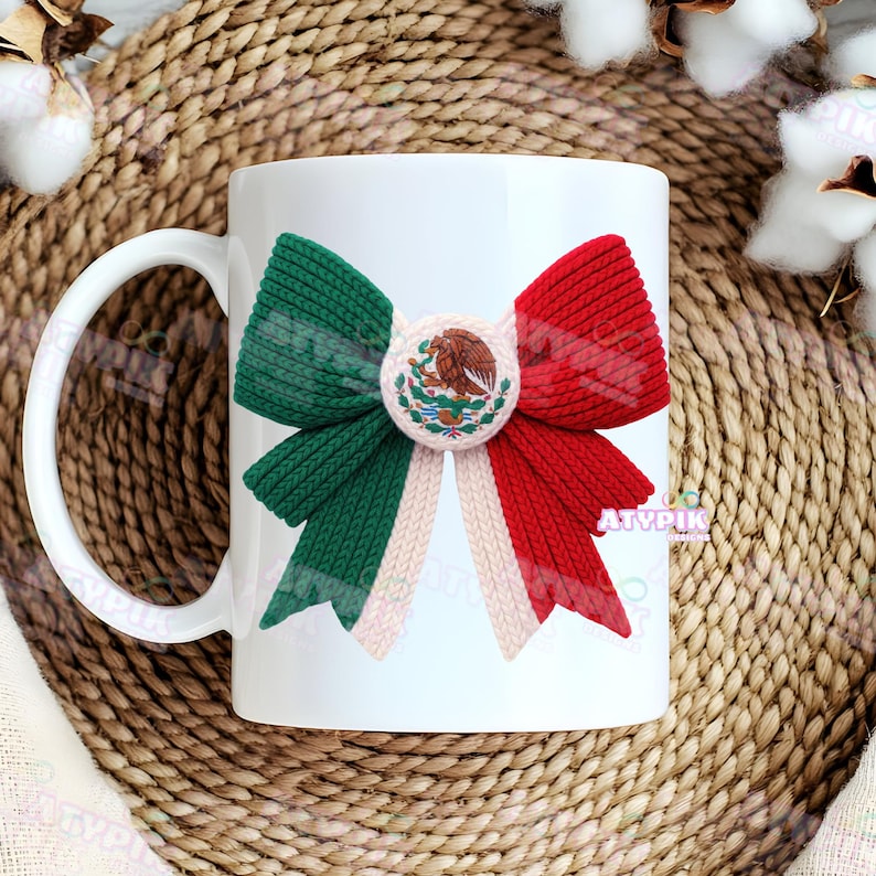 May include: White ceramic mug decorated with a bow in the colours of the Mexican flag: green, white, and red. The bow has a central circular emblem. The mug has a white handle and is set against a woven background.