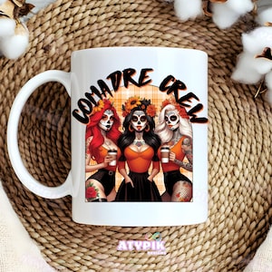 May include: White ceramic coffee mug with a black and orange design featuring three women with sugar skulls and the text "Comadre Crew".