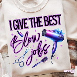 Puede incluir: Sudadera color crema con el texto "I GIVE THE BEST BLOW JOBS" en negro y morado. El diseño incluye un secador de pelo morado y azul, unas tijeras, un peine y un pulverizador. Acentos de estrellas moradas salpican el diseño.
