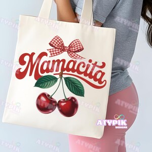 K&ouml;nnte beinhalten: Cremefarbene Tragetasche mit dem Wort "Mamacita" in roter Schrift, einer roten Vichy-Schleife und einem Paar Kirschen. Das Design ist schlicht, mit Fokus auf dem Text und der Fruchtabbildung. Die Tasche hat cremefarbene Griffe.