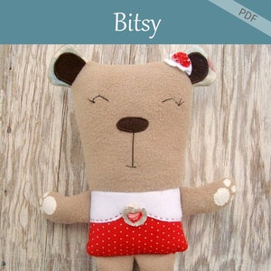 以下が含まれることがあります： 手作りの Bitsy ベアのぬいぐるみ。薄茶色の体、濃い茶色の鼻、赤と白のドット柄のスカートが特徴です。クマの頭には小さな赤い花があり、上部には「Bitsy」の文字があります。Patchwork Posse製です。