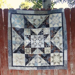 May include: A handmade quilt with a starburst design, featuring a mix of geometric shapes and patterns. The quilt showcases a palette of neutral colors, including cream, gray, and black, with accents of blue and brown. The quilt is displayed outdoors, hanging on a red wooden fence.