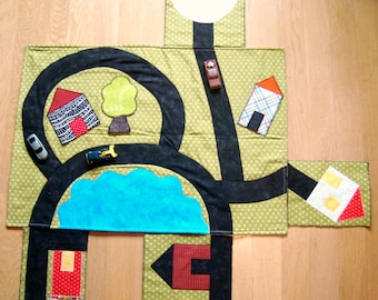 May include: A handmade, green fabric play mat with a road design, featuring black roads, a blue lake, and small fabric houses and trees. Includes toy cars. The mat is designed for children's play and imaginative scenarios.
