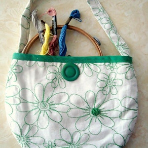 Puede incluir: Bolso tote de tela blanca con un patrón de margaritas bordadas en verde. El bolso tiene un ribete verde y un cierre de botón verde. En el interior, un bastidor de bordado de madera y varios ovillos de hilo de bordar de colores.