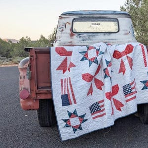 Può includere: Una trapunta bianca con fiocchi rossi, stelle e motivi a bandiera americana drappeggiata sul portellone di un camion rosso vintage. Il design della trapunta include elementi rossi, bianchi e blu, evocando un tema patriottico.