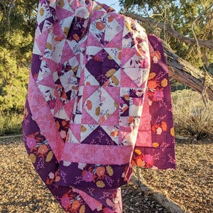 Peut inclure: Une courtepointe faite à la main drapée sur une branche d'arbre. La courtepointe présente un motif patchwork avec des triangles et des carrés roses, violets et blancs. Des motifs floraux orange et jaune accentuent le design. La bordure est violet foncé avec des détails floraux.