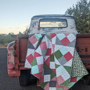 May include: A colorful patchwork quilt draped over the back of a vintage red truck. The quilt features squares of red, green, gray, and white fabric in various patterns. The truck bed is weathered, and the background shows a landscape.
