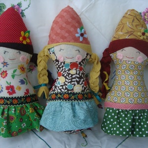 May include: Three handmade fabric dolls with whimsical designs. Each doll has a cone-shaped hat, braided hair, and a smiling face with rosy cheeks. The dolls wear dresses with floral and patterned fabrics, and are decorated with ribbons and flowers.