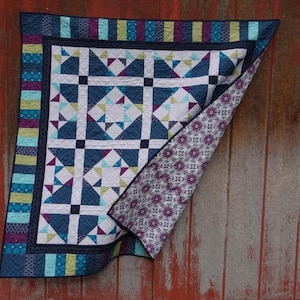 Puede incluir: Un edredón hecho a mano con un diseño geométrico en tonos de azul, blanco, morado y verde. El edredón presenta un patrón repetido de cuadrados y triángulos. El borde tiene un patrón de rayas. El edredón se muestra sobre un fondo de madera rústica teñida de rojo.