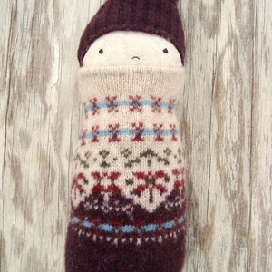 Może przedstawiać: Ręcznie robiona lalka o fantazyjnym designie. Lalka ma bordowy spiczasty kapelusz i kremową twarz z prostymi czarnymi oczami i zakrzywionymi ustami. Ciało to wzorzysta dzianina w kolorze kremowym, bordowym i niebieskim. Lalka jest na drewnianym tle.