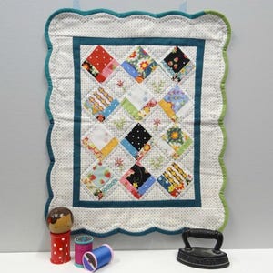 May include: A quilted wall hanging with a patchwork design featuring various colorful fabric squares. The border is scalloped with a teal and green trim. Accessories include a wooden doll, spools of thread, and a small iron.