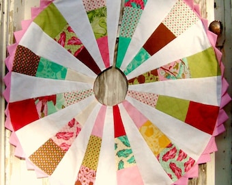 Könnte beinhalten: Ein handgefertigter, kreisförmiger Weihnachtsbaumrock mit Patchwork-Design. Der Rock hat abwechselnd weiße und gemusterte Stoffkeile in verschiedenen Farben, darunter Rosa, Grün, Rot und Braun. Ein rosa gewellter Rand umrahmt den Rock und sorgt für einen festlichen Look.