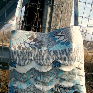 May include: A handmade fabric shoulder bag with a floral pattern in shades of blue, gray, and cream. The bag features a scalloped edge design and a matching patterned strap. The bag is hanging on a fence post.