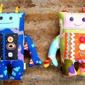 May include: Two handmade robot dolls. One is blue with a black panel and various buttons. The other is green with an orange panel and buttons. Both have button eyes, antennae, and smiling faces.