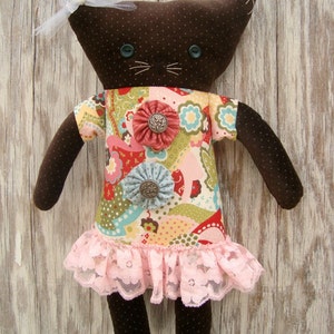 Può includere: Bambola a forma di gatto marrone fatta a mano con un fiocco bianco, indossa un abito floreale colorato con un bordo in pizzo rosa. La bambola ha occhi blu a bottone e baffi ricamati. L'abito è adornato con abbellimenti floreali in tessuto.
