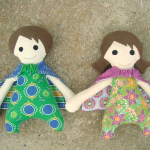 May include: Two handmade fabric dolls with brown hair and smiling faces. One doll wears a green jumpsuit with blue circles, the other a green floral jumpsuit. Both dolls have capes and are holding hands.