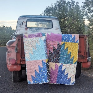 Könnte beinhalten: Zwei farbenfrohe, handgefertigte Quilts mit Sägezahnmuster werden in der Ladefläche eines roten Oldtimer-Pickups präsentiert. Die Quilts weisen verschiedene Muster und Farben auf, darunter Blau, Rosa und Gelb, was eine rustikale und gemütliche Ästhetik erzeugt.