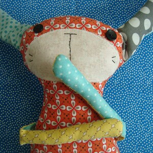 May include: A handmade stuffed bunny doll with orange floral patterned fabric, accented with blue and gray polka dots. The bunny has black button eyes, a stitched nose, and a yellow and green patterned scarf.