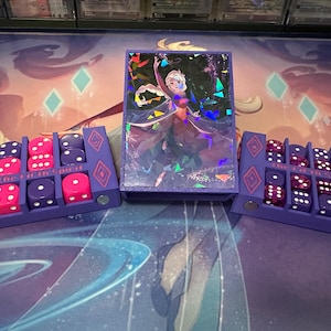 May include: A purple card holder featuring a holographic image of a Frozen character. Two purple dice trays with pink and purple dice flank the card holder. The dice trays are inscribed with the text "The Fifth Spirit".