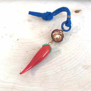 May include: A red glass chili pepper charm hangs from a blue suede cord with a brown and white bead.