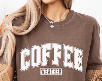 Könnte beinhalten: Ein braunes kurzärmeliges T-Shirt mit dem weißen Schriftzug COFFEE in einer großen College-Schriftart und dem Wort WEATHER in einem kleineren rechteckigen Kasten darunter. Das Shirt wird mit einer beigen Strickjacke und einer schwarzen Leggings getragen.