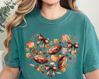 Puede incluir: Camiseta de color verde azulado con un diseño en forma de corazón con balones de fútbol americano, calabazas, lazos y hojas de otoño. El diseño incluye un pequeño suéter, una taza de café y bellotas. La camiseta es de material suave y tiene un corte holgado.