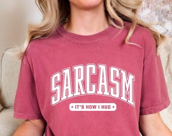 Puede incluir: Una camiseta de manga corta de color rojo oscuro con el texto blanco SARCASM en una fuente universitaria grande y arqueada, con el texto blanco más pequeño IT'S HOW I HUG dentro de un borde rectangular justo debajo de la palabra principal.