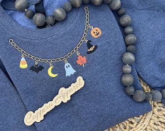 Puede incluir: Una sudadera azul marino jaspeado con un diseño bordado de cadena dorada y dijes colgantes que incluyen un caramelo de maíz, murciélago, luna creciente, fantasma, hoja de arce, sombrero de bruja y calabaza. Un letrero de madera dice Cappora Apparel.