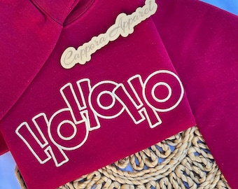 Puede incluir: Una sudadera de manga larga de color rojo oscuro con el texto "HOHOHO" bordado en hilo blanco en el pecho. Una pequeña etiqueta de madera con el texto "Cappora Apparel" en cursiva se encuentra sobre la prenda.