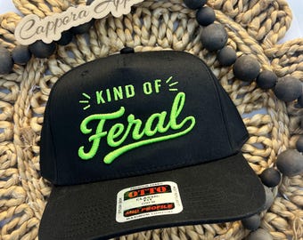 Puede incluir: Una gorra de béisbol negra con el texto bordado "KIND OF Feral" en verde lima brillante. La gorra incluye una pegatina rectangular en la visera con el texto "OTTO PREMIUM FABRIC CLASSIC FIT OSFM MID PROFILE MAXIMUM COMFORT STYLISH LOOK."