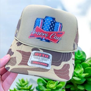 May include: A beige and camouflage trucker hat with a red, white, and blue graphic of three cans of beer with the text "Ameri Can" on the front. The hat has a black mesh back and a white label with the text "OTTO World Famous Foam Front OSFM Trucker Durable Snap Closure".