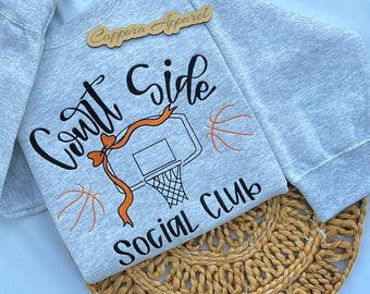 Puede incluir: Una sudadera gris claro de cuello redondo con el texto en cursiva negra Court Side Social Club. El diseño incluye una canasta de baloncesto bordada, dos balones de baloncesto naranjas y una cinta naranja con un lazo.
