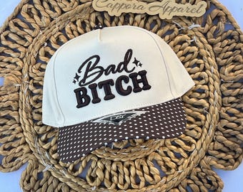 Puede incluir: Una gorra de béisbol color crema con el texto bordado "Bad BITCH" en marrón oscuro y pequeños detalles de estrellas. La visera es marrón oscuro con un patrón de lunares blancos e incluye una pequeña etiqueta de la marca D&Y al frente.