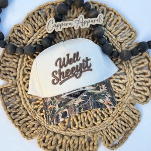May include: A white baseball cap with a brown embroidered text that reads Well SHEEYIT. The brim features a camouflage pattern with a David & Young D&Y sticker. The cap is positioned on a woven circular mat surrounded by dark wooden beads.