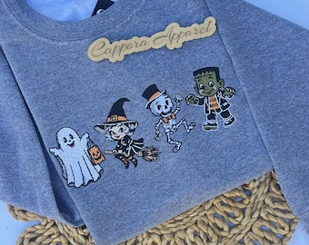 Puede incluir: Una sudadera gris jaspeado de cuello redondo con un diseño bordado de un fantasma blanco, una bruja en una escoba, un esqueleto con sombrero de copa y un monstruo verde. El texto Cappora Apparel está bordado en color dorado sobre los personajes.