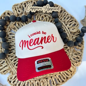 May include: A white and red baseball cap with the embroidered phrase "i could be meaner." The cap has a red brim and a white crown. The hat is a classic fit, mid-profile, and made of premium fabric.