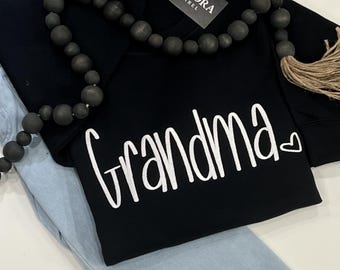 Puede incluir: Una camiseta negra de manga larga con el texto blanco "grandma" en letra cursiva y un pequeño corazón al final. Una etiqueta negra con el texto "CAPPORA APPAREL" está unida al cuello y un collar de cuentas de madera oscura se extiende sobre ella.