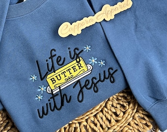 Puede incluir: Una sudadera de cuello redondo en color azul polvoriento con texto bordado en negro que dice Life is with Jesus, junto a un gráfico de una barra de mantequilla amarilla. Pequeñas flores bordadas en azul claro rodean el texto. Un letrero de madera dice Cappora Apparel.