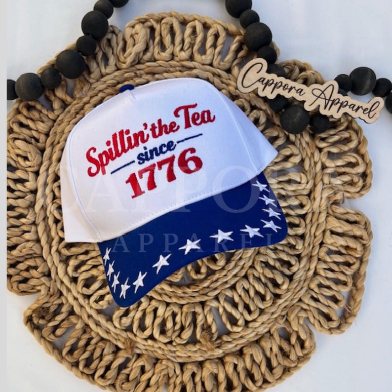 K&ouml;nnte beinhalten: Eine wei&szlig;e Baseballkappe mit blauem Schirm, der mit wei&szlig;en Sternen verziert ist. Auf der Kappe ist der Text "Spillin' the Tea since 1776" in Rot und Blau gestickt. Der Hut wird auf einem gewebten, kreisf&ouml;rmigen Platzdeckchen pr&auml;sentiert.