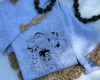 Op de afbeelding: Een lichtgrijs sweatshirt met een zwarte lijntekening van Winnie de Poeh en zijn vrienden. Het sweatshirt heeft lange mouwen en de tekst "Cappera Apparel" op een houten label.