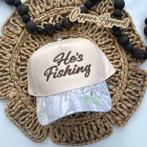 Puede incluir: Gorra de b&eacute;isbol beige con el texto bordado "He's Fishing" en escritura marr&oacute;n. La visera de la gorra presenta un estampado de camuflaje en tonos grises y verdes. La gorra se exhibe sobre un salvamanteles circular tejido.