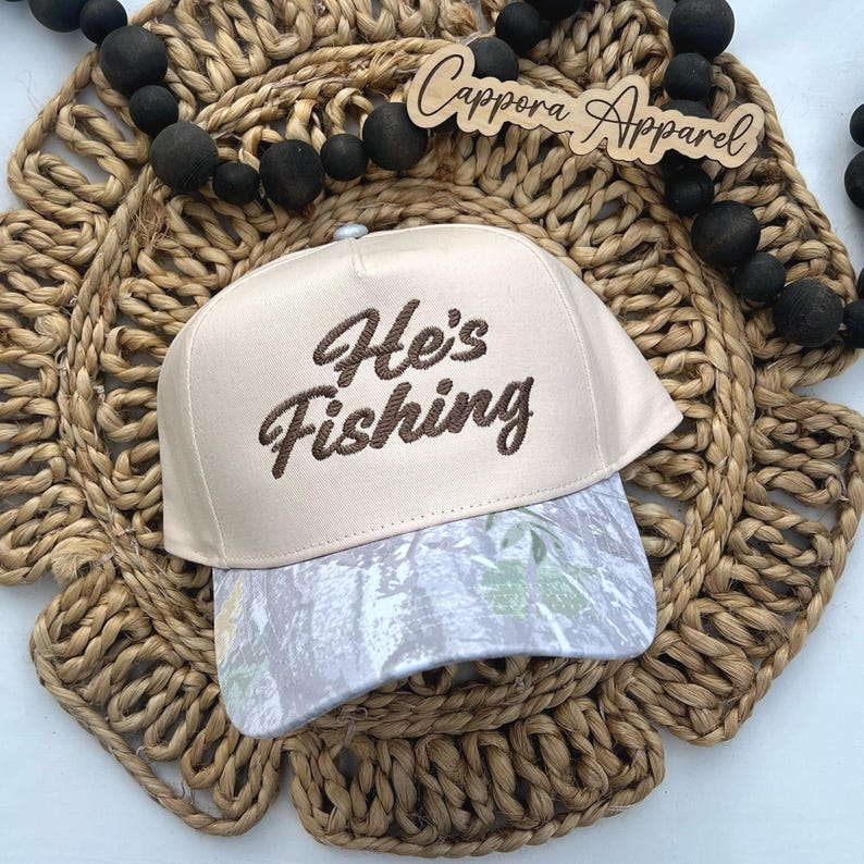 Puede incluir: Gorra de b&eacute;isbol beige con el texto bordado "He's Fishing" en escritura marr&oacute;n. La visera de la gorra presenta un estampado de camuflaje en tonos grises y verdes. La gorra se exhibe sobre un salvamanteles circular tejido.