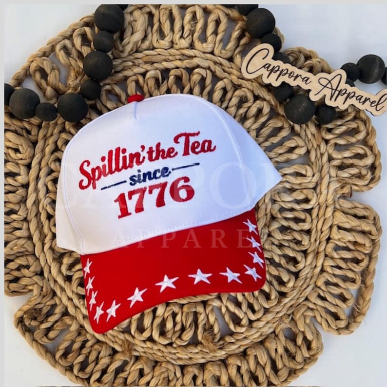 K&ouml;nnte beinhalten: Eine wei&szlig;-rote Baseballkappe mit dem Text "Spillin' the Tea since 1776". Der rote Schirm ist mit wei&szlig;en Sternen verziert. Die Kappe wird auf einem gewebten, kreisf&ouml;rmigen Untersetzer pr&auml;sentiert.
