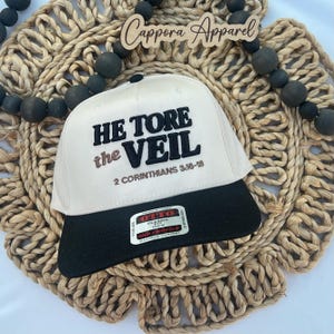 Puede incluir: Gorra de béisbol beige y negra con el texto "HE TORE the VEIL 2 CORINTHIANS 3:16-18" en letras negras y marrones. La gorra tiene una visera negra y una etiqueta blanca.