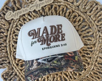 May include: A cream-coloured baseball cap featuring the embroidered brown text MADE for MORE and EPHESIANS 2:10 on the front panel. The brim displays a camouflage pattern in shades of brown, green, and black, with a D&Y brand sticker attached.