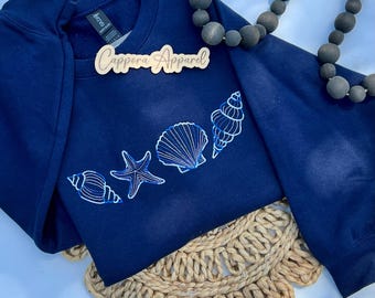 May include: Navy blue sweatshirt with embroidered seashells and a starfish design. The text "Cappora Apparel" is on a wooden tag. The sweatshirt is displayed on a woven placemat with a beaded necklace.
