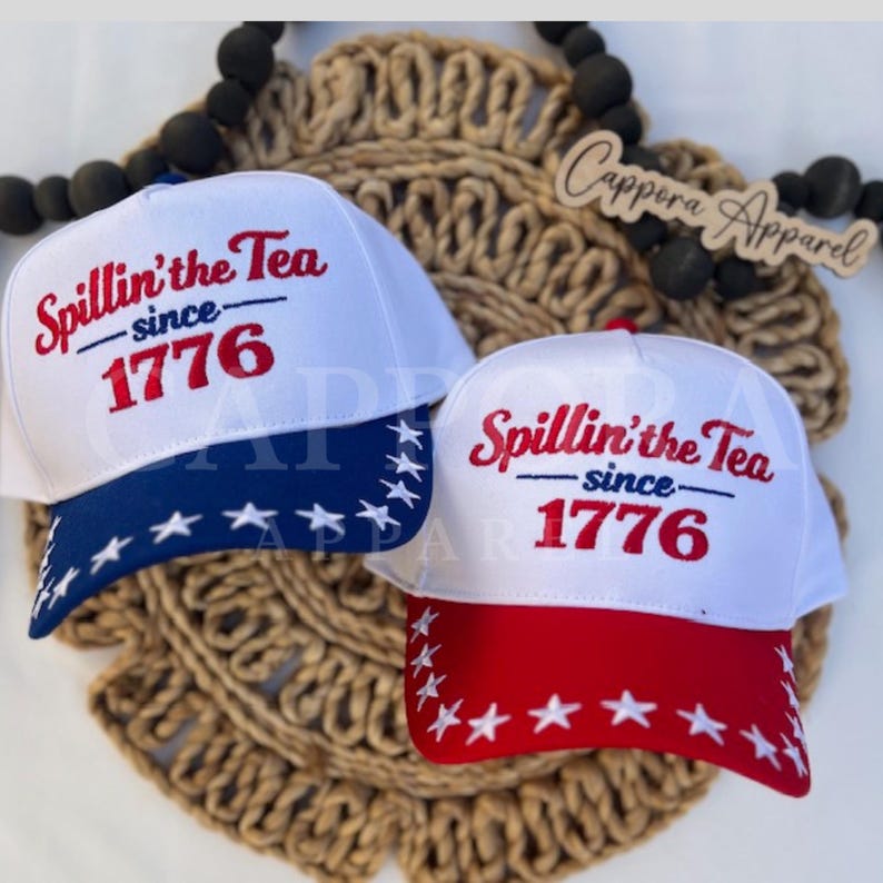 K&ouml;nnte beinhalten: Zwei wei&szlig;e Baseballkappen mit dem Text "Spillin' the Tea since 1776" in Rot gestickt. Eine Kappe hat einen blauen Schirm mit wei&szlig;en Sternen, die andere einen roten Schirm mit wei&szlig;en Sternen. Die Kappen liegen auf einer gewebten, kreisf&ouml;rmigen Matte.